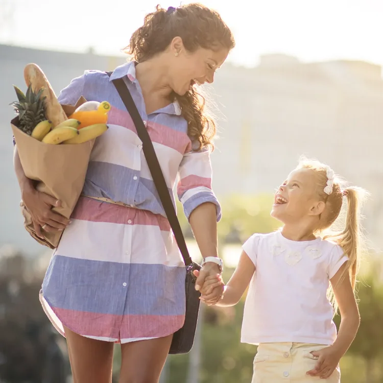 Junge Frau und Mädchen laufen Hand in Hand, Frau mit Lebensmitteln in einer Tüte unter dem Arm - Familienmanager:in