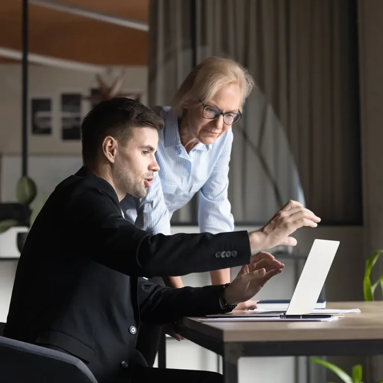 Junger Mann sitzt am Tisch mit Laptop, eine Frau steht daneben und schaut mit auf den Bildschirm - Personal Assistant